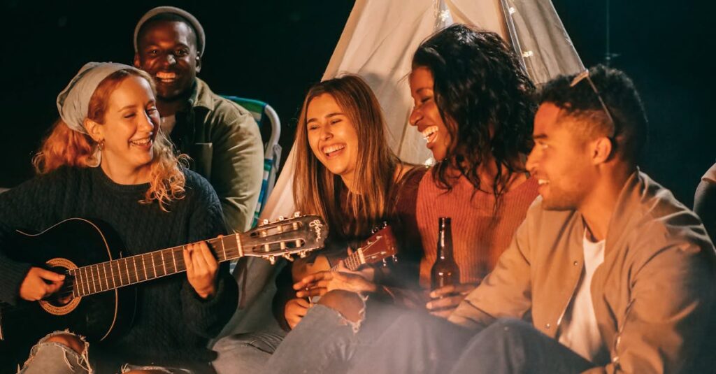 A diverse group of friends playing guitar and ukulele by a campfire at night, having fun and socializing.